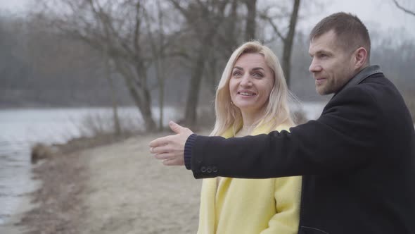 Positive Caucasian Man Opening Eyes of Blond Smiling Woman on Riverbank and Kissing Sweetheart