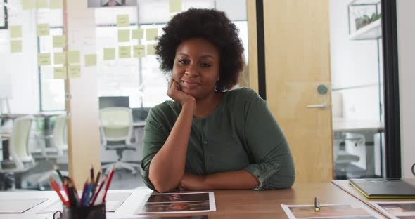 Portrait of african american businesswoman sitting at desk leaning on hand and smiling in office