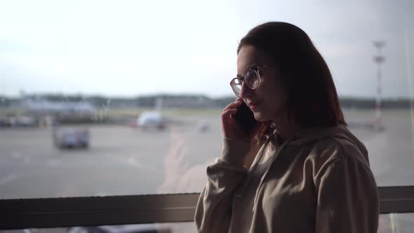 A Young Woman in a Sweatshirt and Glasses Speaks on the Phone Against the Background of a Window at