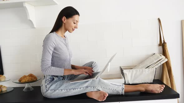 Image of Cute Brunette Woman Sitting at Kitchen and Using Laptop