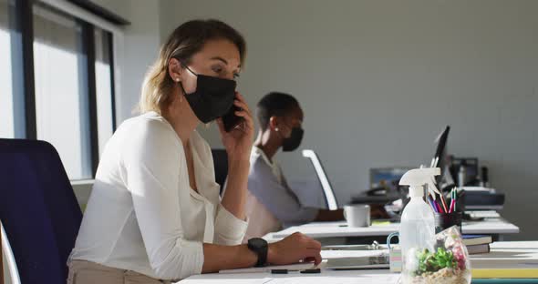 Two diverse female colleagues wearing face mask, working at desk in office