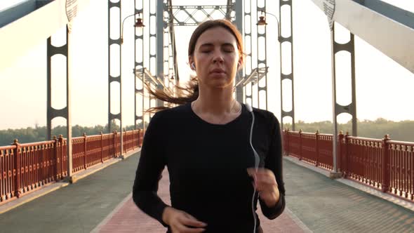 Slow Motion Scene of an Athletic Girl in Black Sportswear Runs Across the Bridge During Dawn Towards