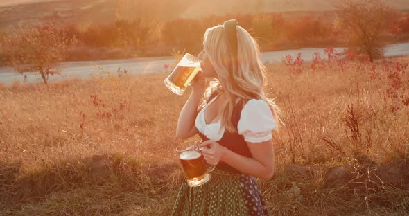 Joyful Slavic Girl in Dress Holds Two Pints of Beer Drinks and Smiles on Nature