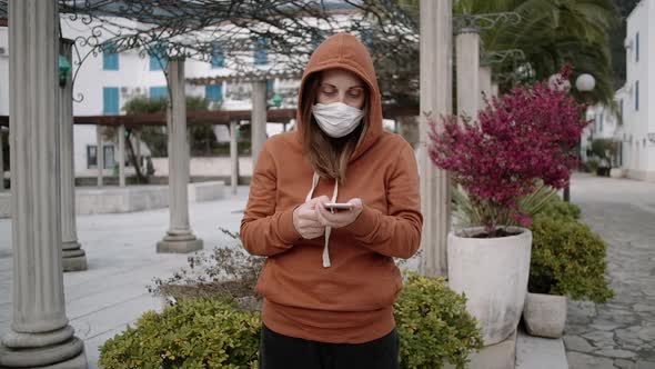 Woman in orange sweater and medical mask using her phone on empty street.