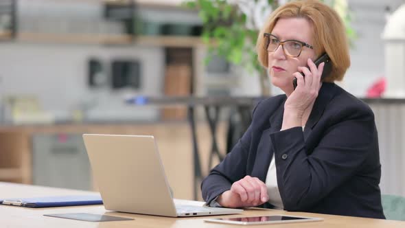 Old Businesswoman with Laptop Talking on Smartphone in Office