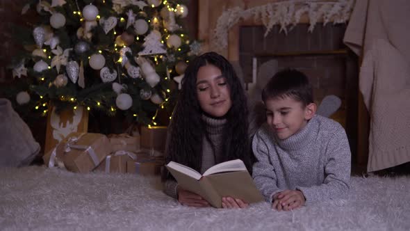 Mom Reads a Book To Her Son and Kisses Him Lying on the Floor Near the Christmas Tree on Christmas