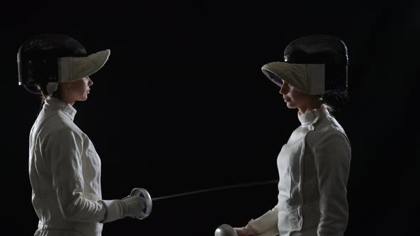 Side View of Two Young Female Swordsmen Saluting with Rapiers and Putting on Helmet