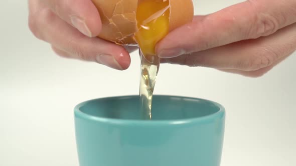Bright yellow egg yolk flows from a broken eggshell in the chef hands into a blue ceramic mug. 