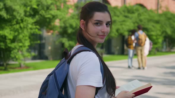 Follow Shot of Happy University Student Walking with Book and Looking Back at Camera Outdoors