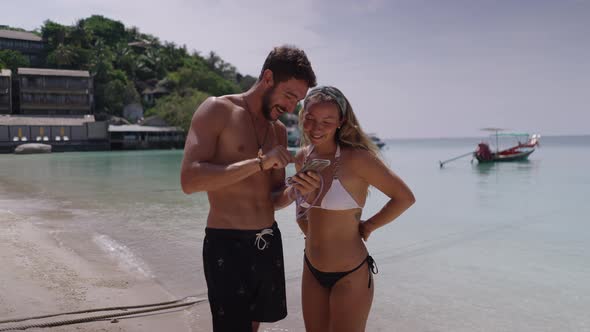 A Static Shot of a Young Couple Looking at Smartphone While Standing on a Beach in a Sunny Day