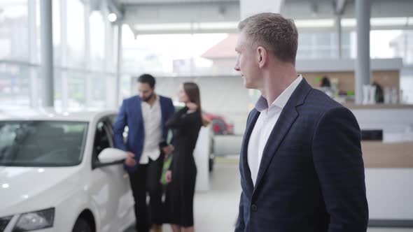 Confident Caucasian Car Dealer Looking Back at Happy Multiracial Couple Opening New White Car and