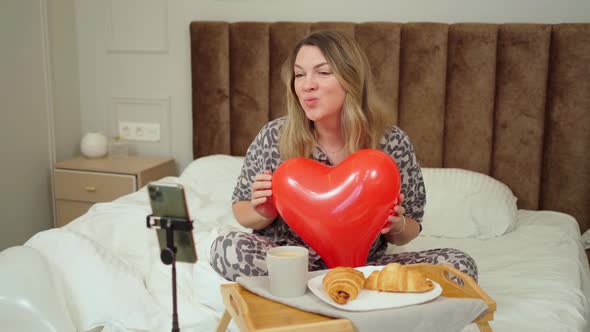 Woman Sits in Bed with a Heartshaped Balloon and Breakfast on the Table There is a Phone on a Tripod