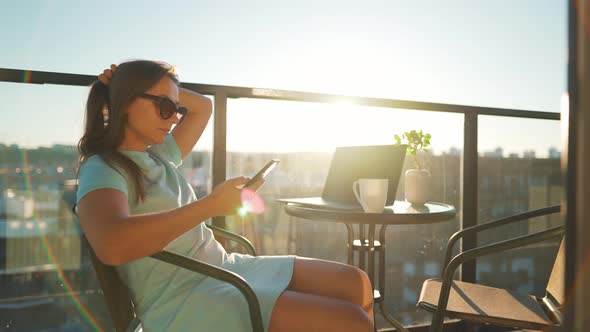 Woman Using Smartphone on the Balcony Against the Backdrop of the Sunset. Remote Work Concept