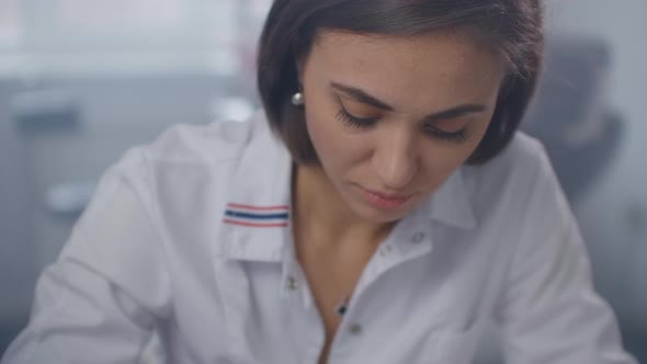 A Female Doctor Sits at a Table and Writes with a Pen Writing Out a Prescription To a Patient