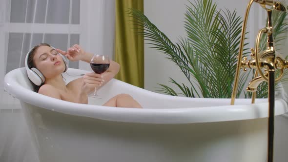 Beautiful Young Woman Enjoying Pleasant Bath with Foam, Holding a Glass of Wine, Woman Listening To Music