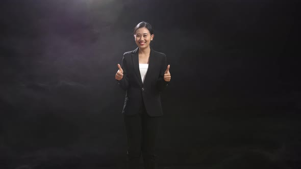 Smiling Asian Speaker Woman In Business Suit Showing Thumbs Up Gesture To Camera In The Black Studio