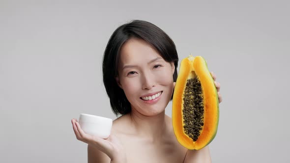 Mature Japanese Woman Holding Papaya And Moisturizer Over Gray Background