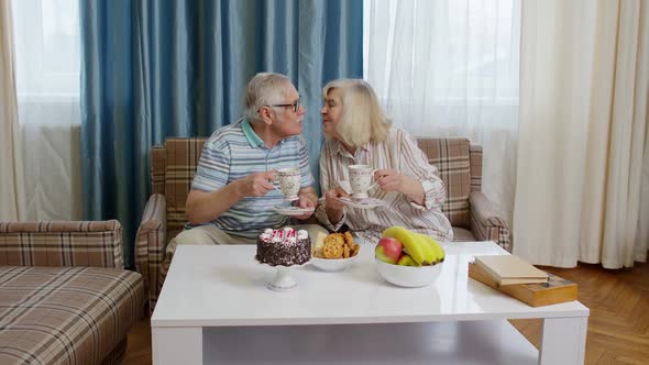 Senior Couple Holding Drinking Tea Talking Laughing Smiling Kissing Sitting on Sofa at Home