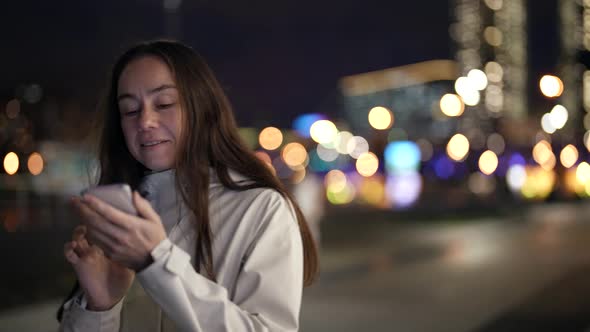 a Woman with Long Hair and a Light Jacket is Typing a Message on Phone and Laughing in the Evening