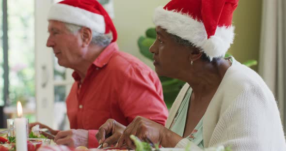 Diverse senior man and woman in santa hats listening and eating at christmas dinner table at home