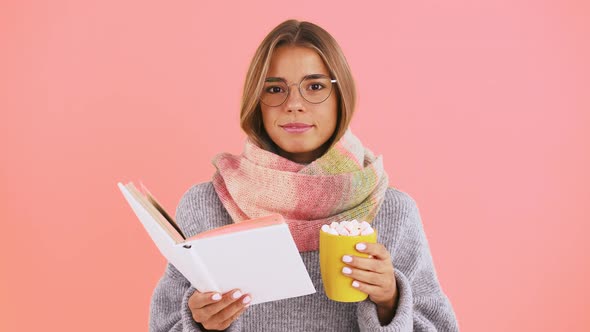 Lady in Sweater and Scarf is Reading a Book Holding Yellow Cup of Coffee Decorated By Marshmallow