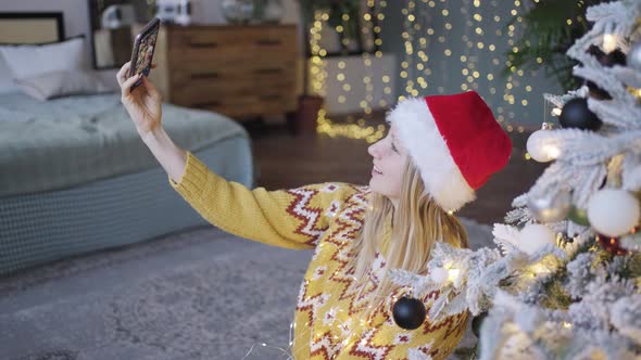 Cute Woman Takes a Selfie on Her Phone Against the Background of a Christmas Tree