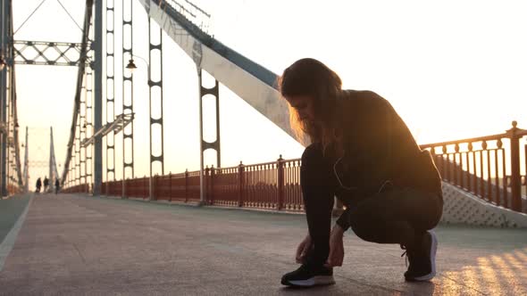 Athletic Girl Tying Shoelaces on Sneakers and Begins To Run on the Bridge During Dawn. Golden Hour