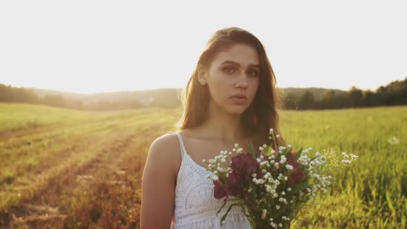 Girl Standing in Meadow with Bouquet of Wild Flowers at Sunset on Summer Evening, Look at Camera