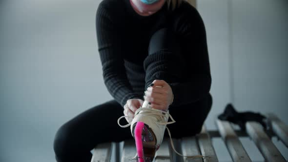 Ice Skating  Young Woman in Medicine Mask Ties Shoelaces on White Skates in Locker Room
