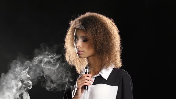 African American Girl Smokes an E-cigarette in an Empty Room. Black Background