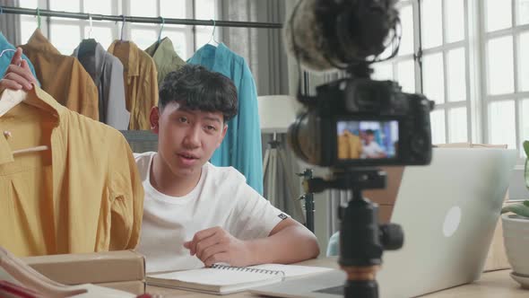 Asian Young Man Looking At Computer And Recording Video By Camera For Selling Clothes Online At Home