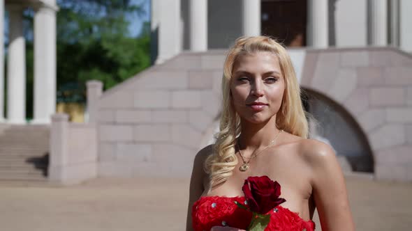 Portrait of a Smiling Blonde with Big Breasts and in a Red Dress with a Rose in Hands Against the