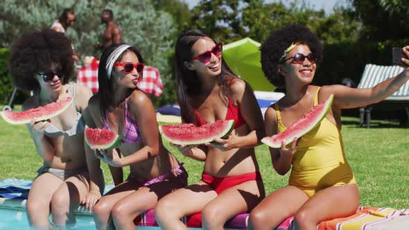 Group of diverse girls holding watermelon taking a selfie together on smartphone while sitting by th