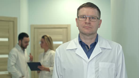 Male Doctor Looking at Camera While Medical Staff Working on the Background