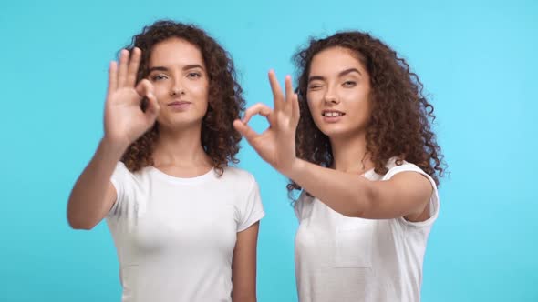 Two Beautiful Female Curly Caucasian Twins in White Tshirts Synchronously Showing Ok on Blue
