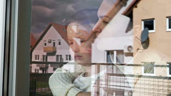 Mother and Little Daughter Stand Embracing and Look Sadly Out the Window