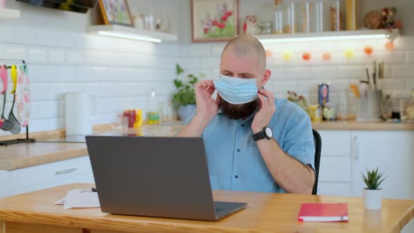 Mature Bearded Man Puts on a Protective Medical Mask While Working or Studying Remotely at Home