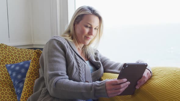 Happy caucasian mature woman looking through the window and using tablet