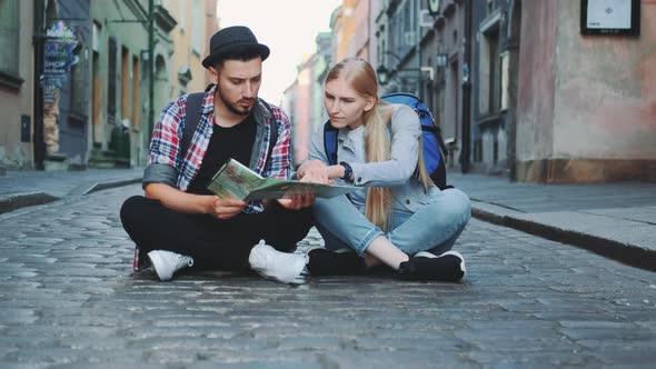 Young Tourists Couple Using Map, Sitting on Pavement and Admiring Historical Surroundings