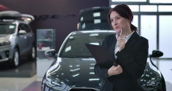 Confident Caucasian Saleswoman Looking Around in Car Dealership and Writing Down in Documents