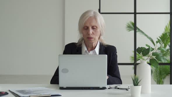 Elderly Business Woman Works in an Office with a Laptop and Looks at the Camera