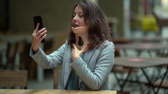 Woman with Orthopedic Collar on Neck Is Communicating By Video Call on Mobile Phone in Cafe, Curing