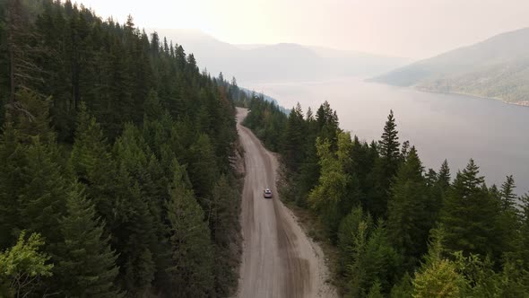 Silver car driving along a dusty forest service road during wildfire season with Adams Lake in the b