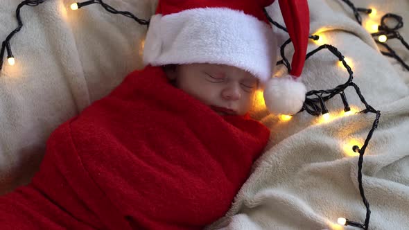 Top View Portrait First Days Of Life Newborn Cute Funny Sleeping Baby In Santa Hat Wrapped In Red