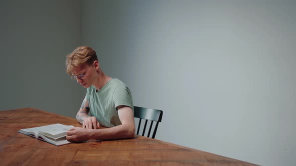 Guy in Glasses Studying By Reading a Textbook Sitting in Empty Grey Room