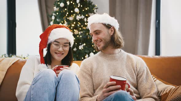 Mixed Race Pair Smiling Cuddling and Holding Red Mugs of Tea Sitting on Sofa at Home Against