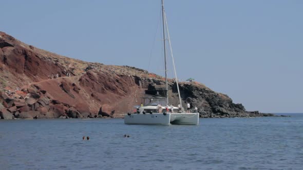 A catamaran sails by the red beach of Santorini.