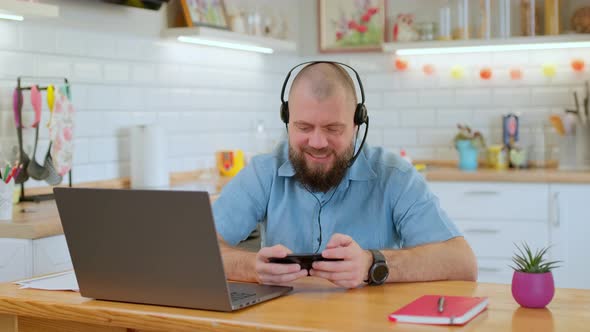 Bearded Mature Man Emotionally Playing a Game on His Phone During Online Conference While Sitting at