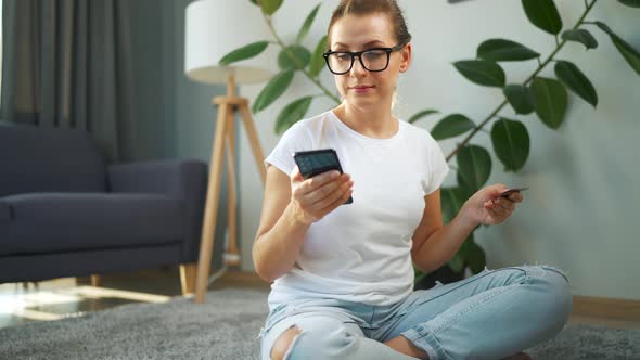 Woman with Glasses is Sitting on the Floor and Makes an Online Purchase Using a Credit Card and