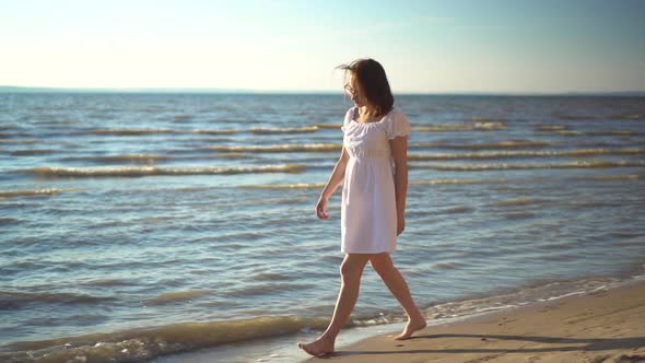 Attractive Young Woman Walks Along the Sea Beach. A Girl in a White Dress Walks Barefoot Along the